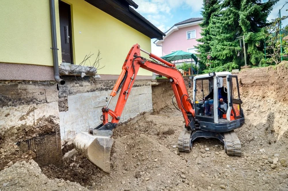 Compact orange excavator digging a foundation trench beside an exposed basement wall at a Toronto home
